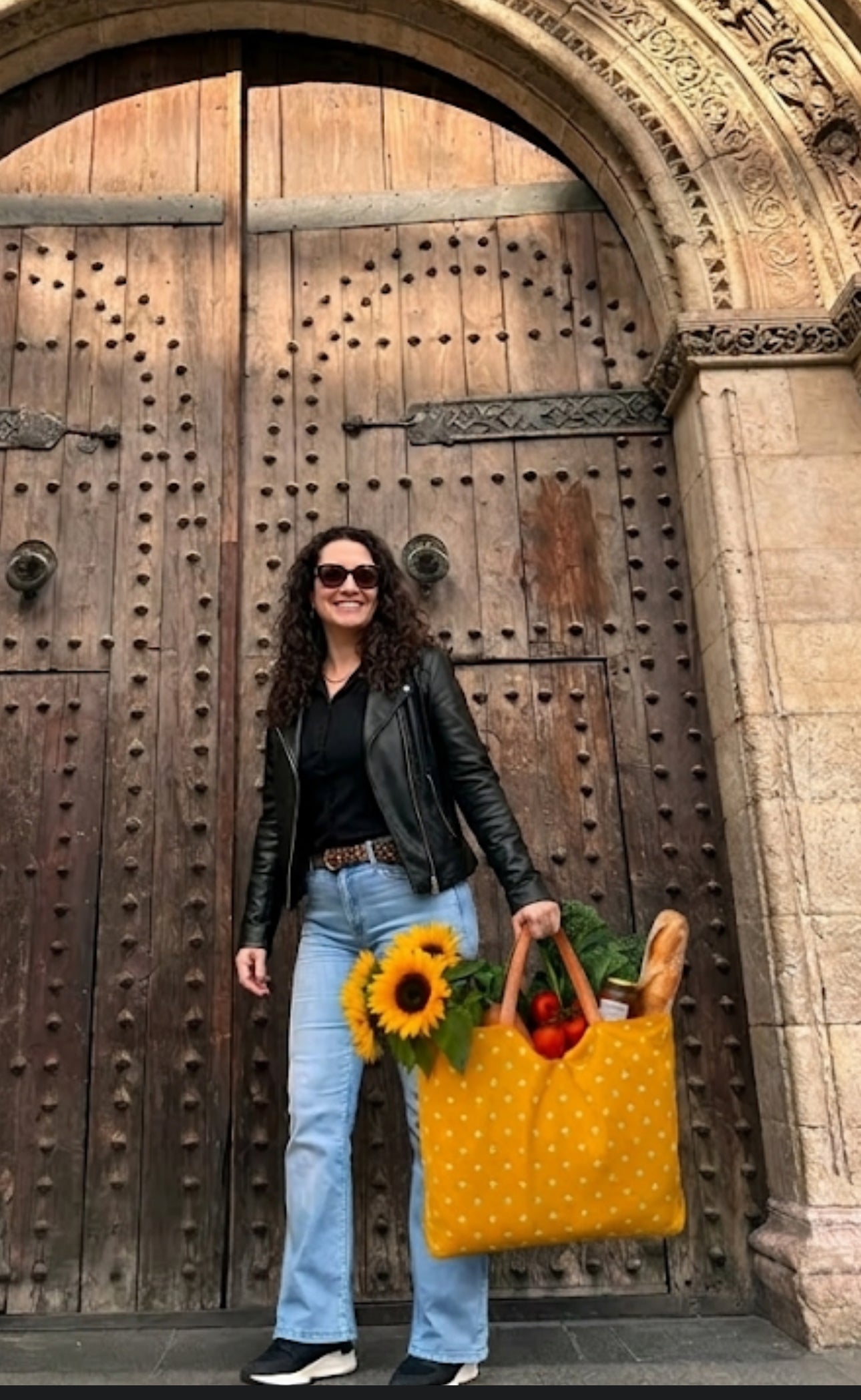 The founder of The Silky Way Studio holding the "Honeycomb" yellow polka dot bag in front of a rustic wooden door. The oversized bag is filled with sunflowers, vegetables, and a baguette, showcasing its large capacity and use as a stylish market tote